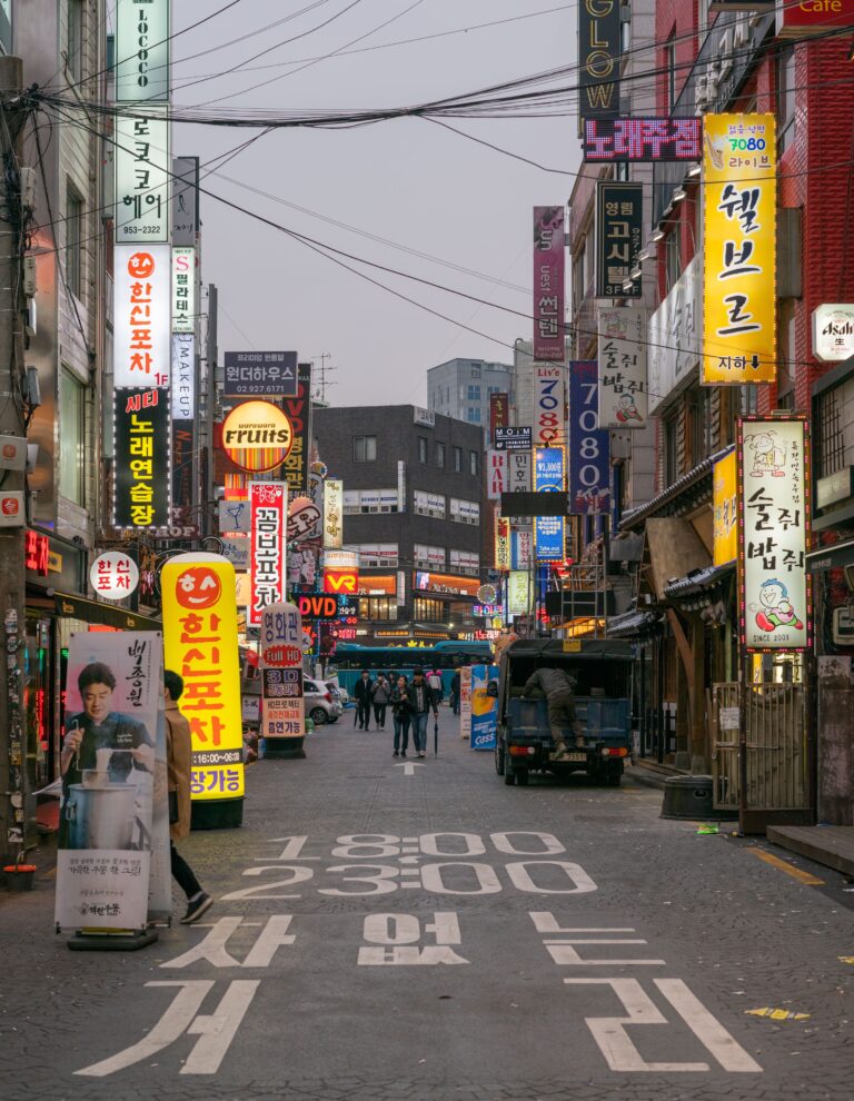Pedestrians walking along a busy shopping street in Seoul, South Korea, lined with Korean shop signs and restaurants.
