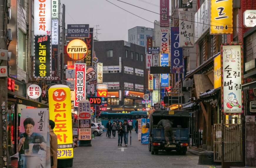 Pedestrians walking along a busy shopping street in Seoul, South Korea, lined with Korean shop signs and restaurants.